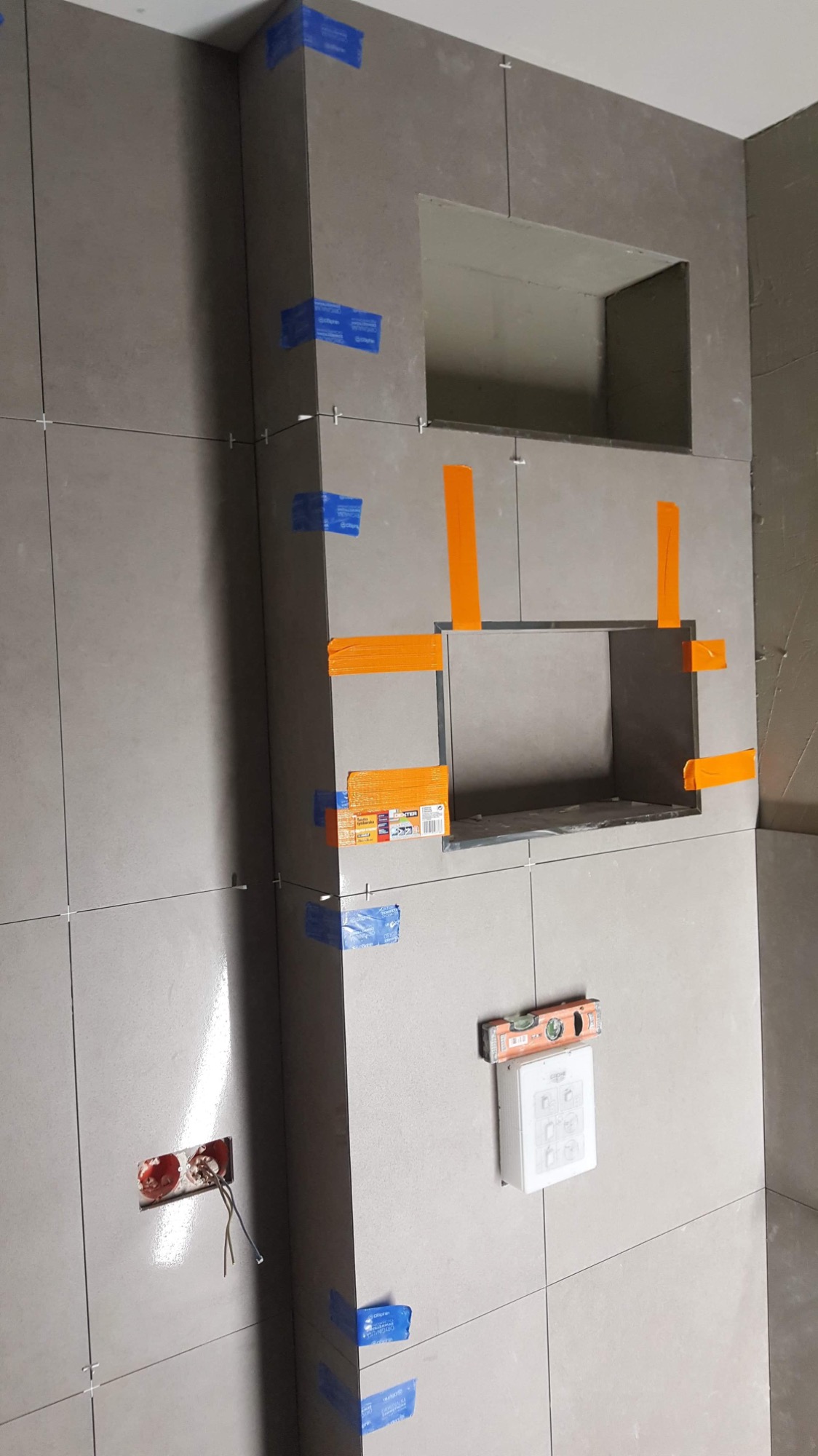 Ściana w trakcie układania płytek ceramicznych w łazience, widoczne wnęki, poziomica, puszka elektryczna z wystającymi przewodami, niebieska taśma maskująca i krzyżyki dystansowe.