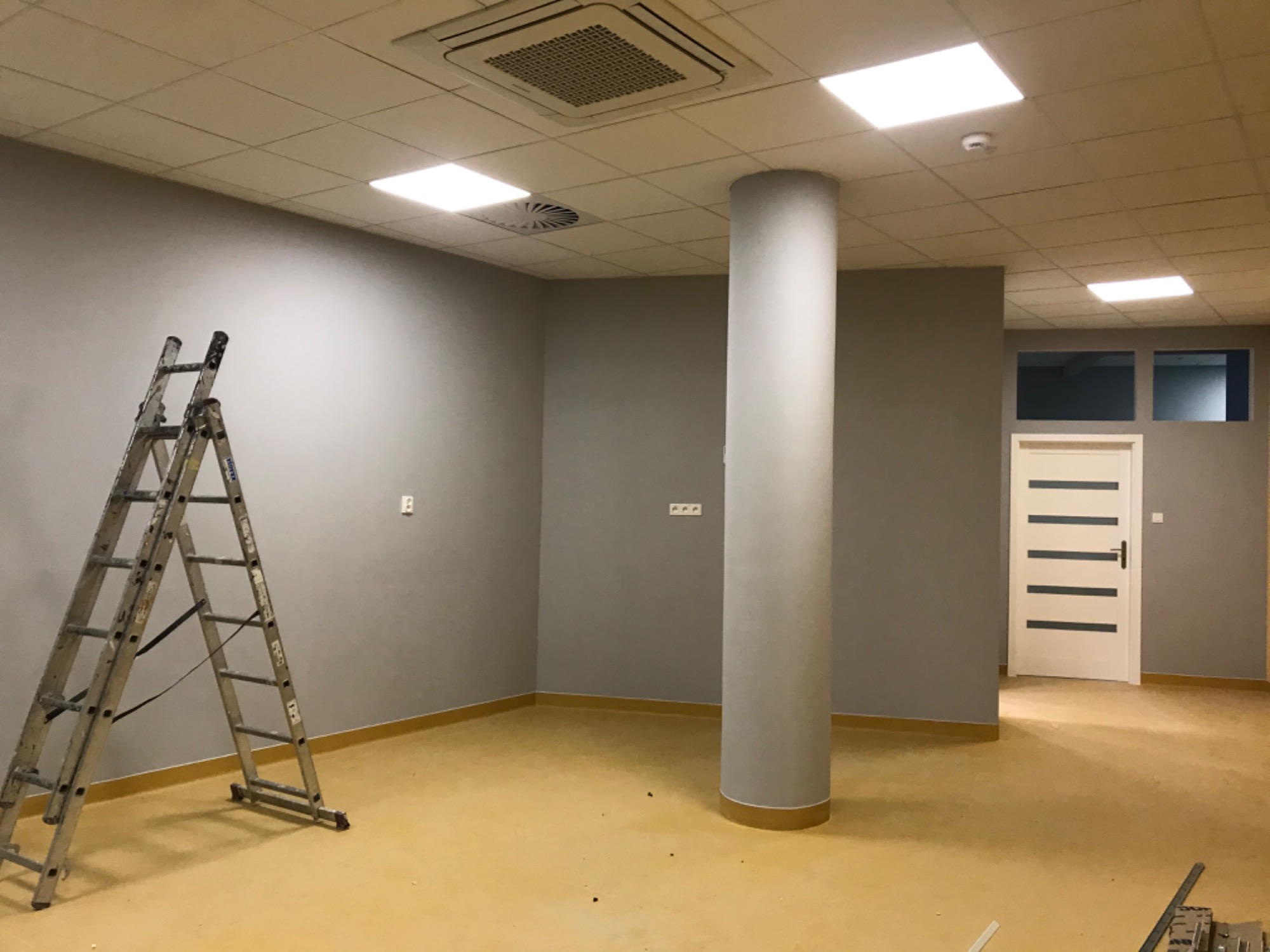 Szare wnętrze biurowe z kolumną, drabina oparta o ścianę, widoczne instalacje sufitowe i drzwi z poziomymi przeszkleniami, podłoga w odcieniach żółci.