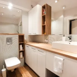 Jasna łazienka z drewnianym blatem, białą umywalką nablatową, mozaiką na ścianach, podwieszaną toaletą i drewnianą podłogą.