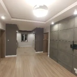 Nowoczesne wnętrze mieszkania po remoncie: panele ścienne imitujące beton, drewnopodobna podłoga, minimalistyczne oświetlenie sufitowe i uchwyt do telewizora na ścianie.