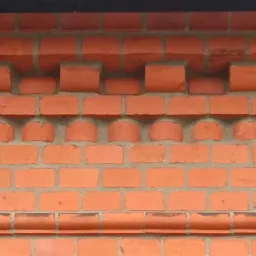 Fragment ściany z czerwonej cegły z dekoracyjnym układem cegieł w górnej części, tworzących wypukłe i wklęsłe wzory.