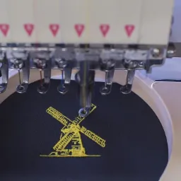 Proces haftowania żółtego wiatraka na ciemnym materiale przy użyciu przemysłowej maszyny do haftowania.