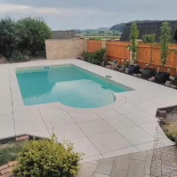 Nowoczesny basen ogrodowy z jasną, kwadratową nawierzchnią z płytek, otoczony zielenią i drewnianym ogrodzeniem, z widokiem na pagórkowaty krajobraz w tle.