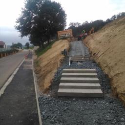 Karol - Budowa schodów zewnętrznych z prefabrykatów betonowych na zboczu, widoczne przygotowane podłoże z kruszywa, w tle maszyny budowlane i tablica reklamowa firmy.