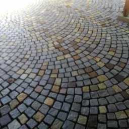 Półokrągła nawierzchnia ułożona z kostki granitowej w odcieniach szarości, beżu i brązu, z widocznym fragmentem drewnianej konstrukcji w tle.