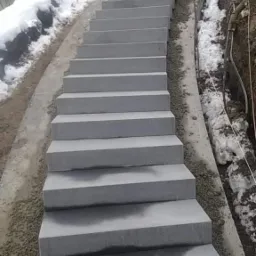 Zewnętrzne schody z szarego kamienia prowadzące pod górę w kierunku domu, częściowo pokryte śniegiem.