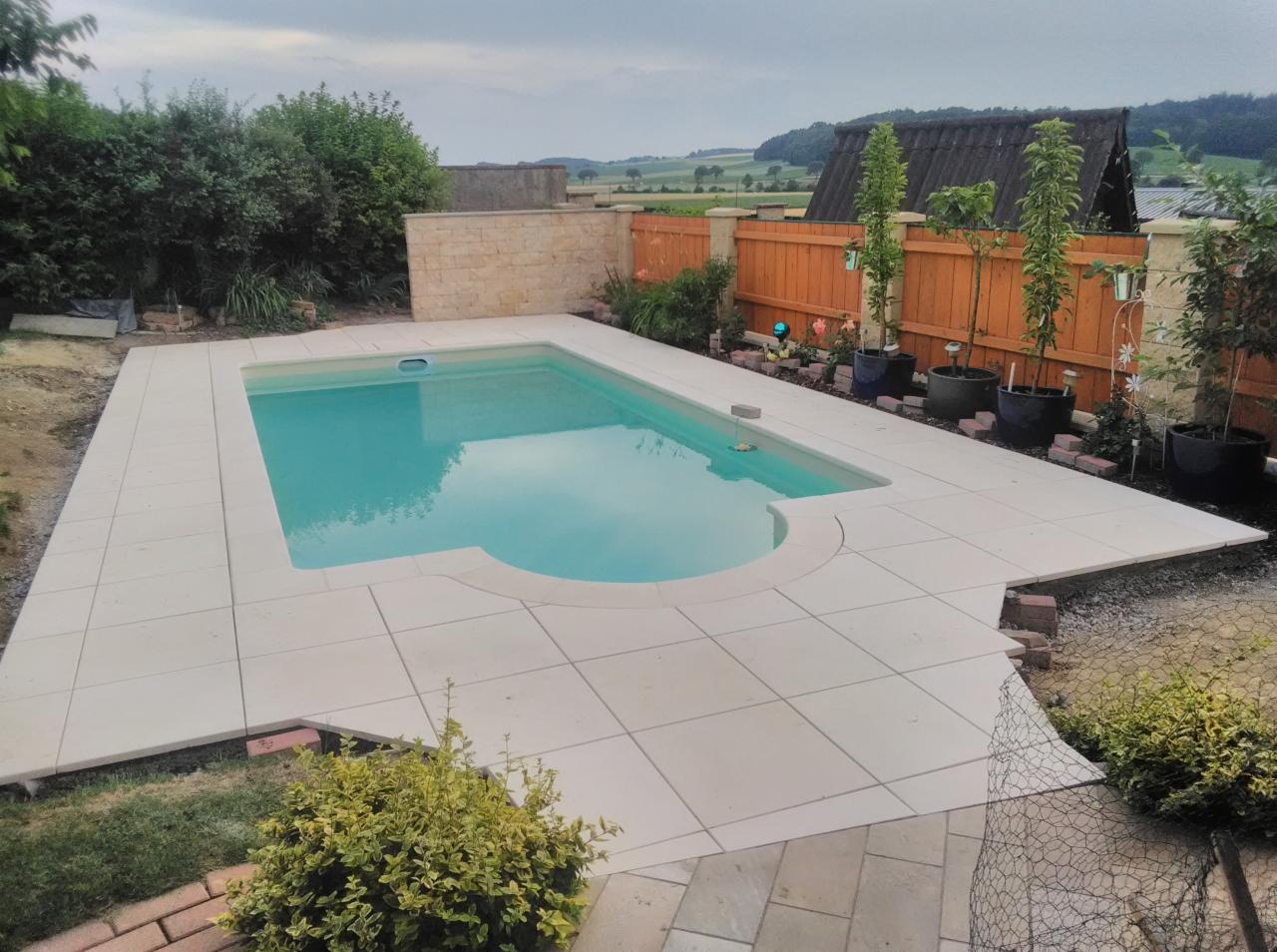 Nowoczesny basen ogrodowy z jasną, kwadratową nawierzchnią z płytek, otoczony zielenią i drewnianym ogrodzeniem, z widokiem na pagórkowaty krajobraz w tle.