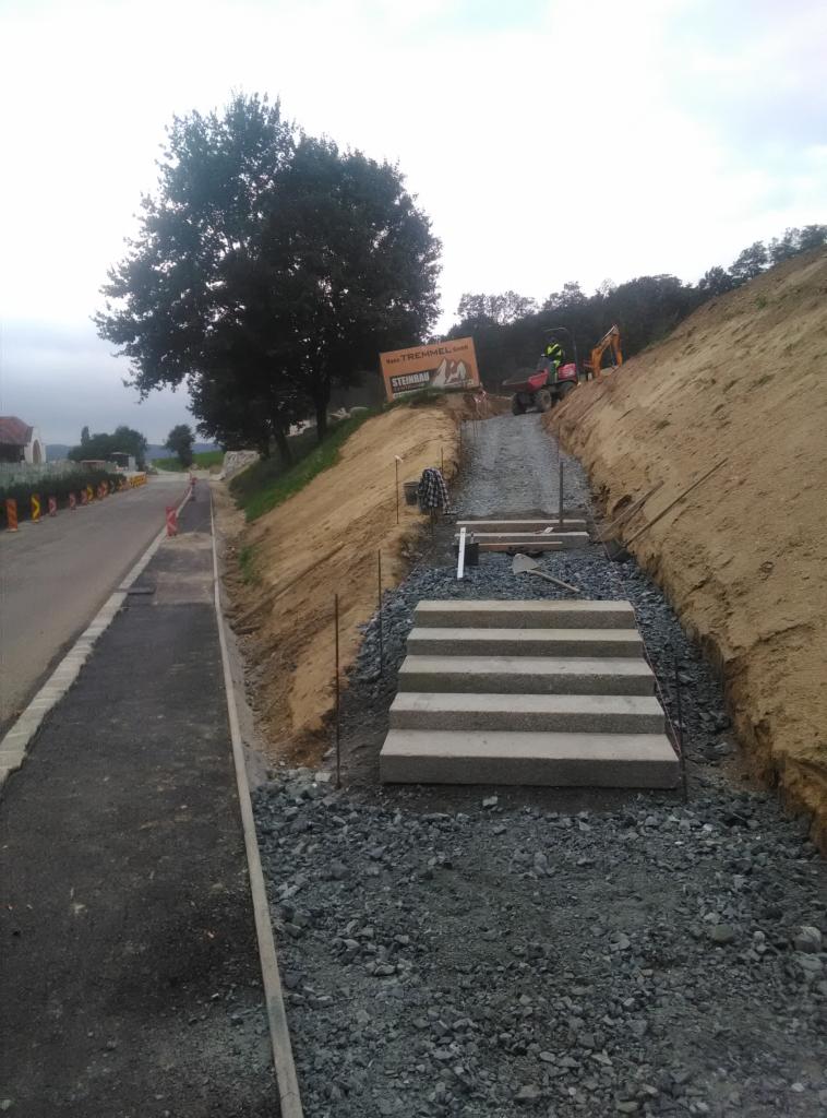 Budowa schodów zewnętrznych z prefabrykatów betonowych na zboczu, widoczne przygotowane podłoże z kruszywa, w tle maszyny budowlane i tablica reklamowa firmy.