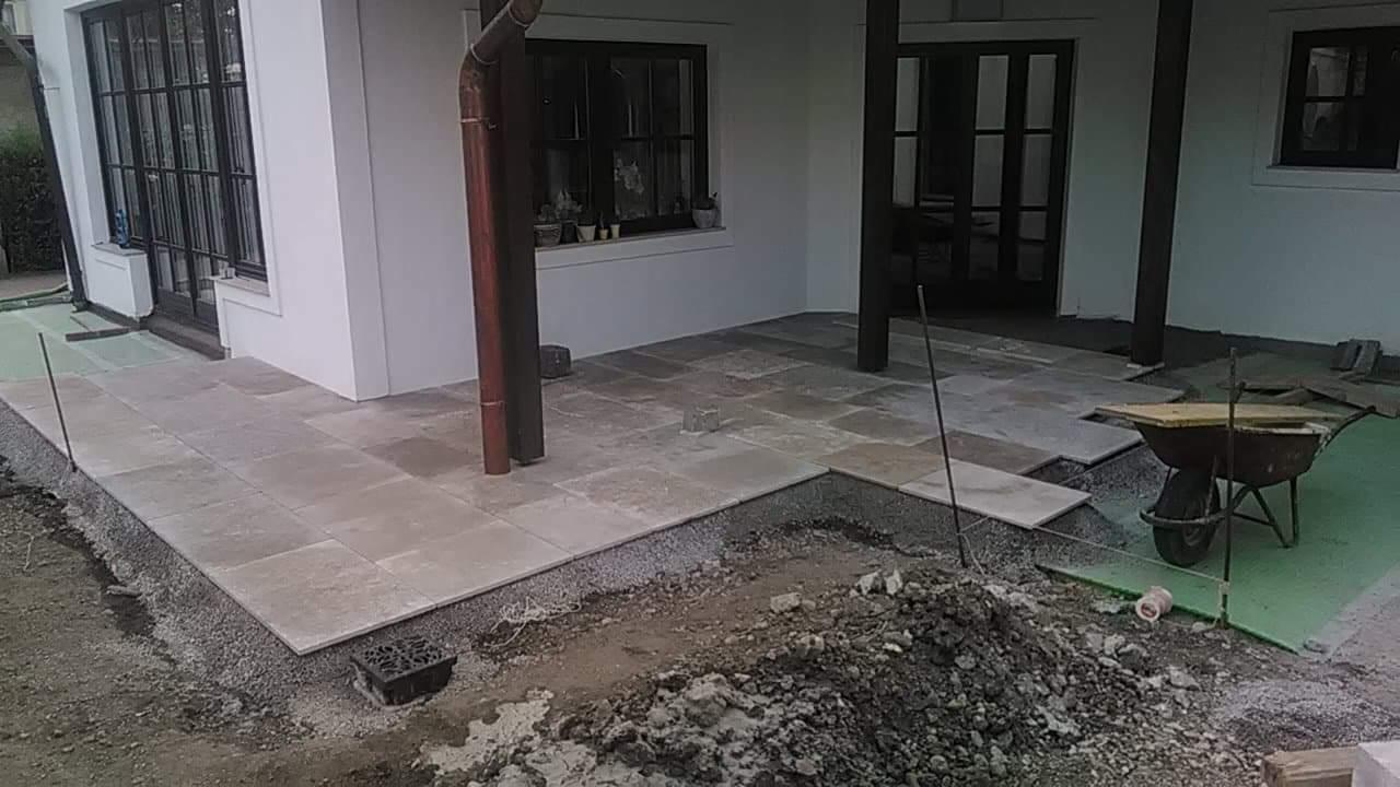 Montaż kamiennego tarasu w Swiebodzicach: widok na częściowo ułożone płyty, przygotowane podłoże z gruzu i piasku, taczka z narzędziami w tle.