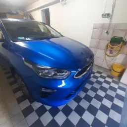 Błękitny samochód Kia zaparkowany w garażu z szachownicową podłogą, widoczny wąż do mycia i żółte wiadro.