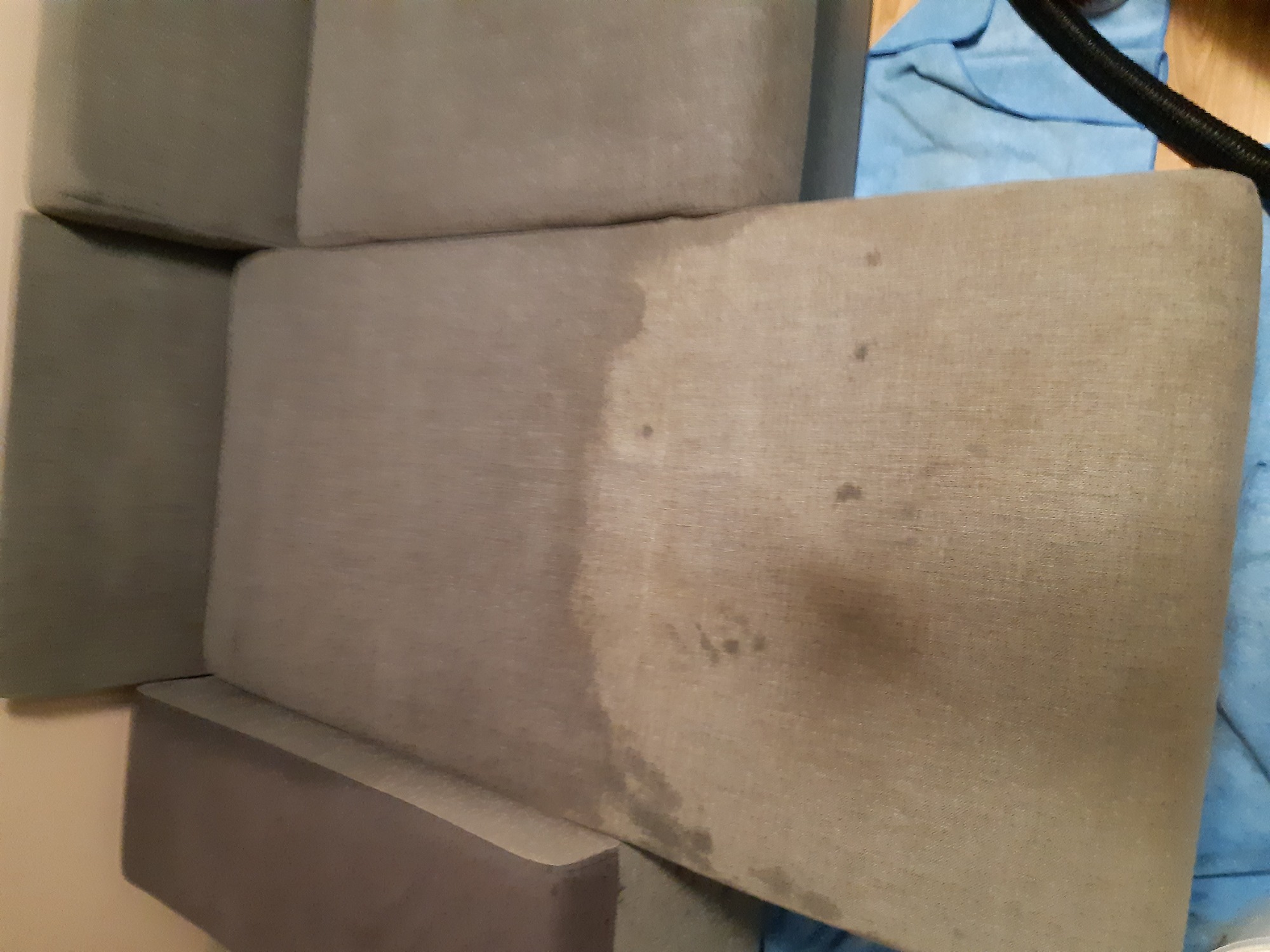 Szary narożnik z widoczną różnicą w czystości tapicerki, część po czyszczeniu jest jaśniejsza i bez plam, obok leży niebieska ściereczka i wąż od odkurzacza.