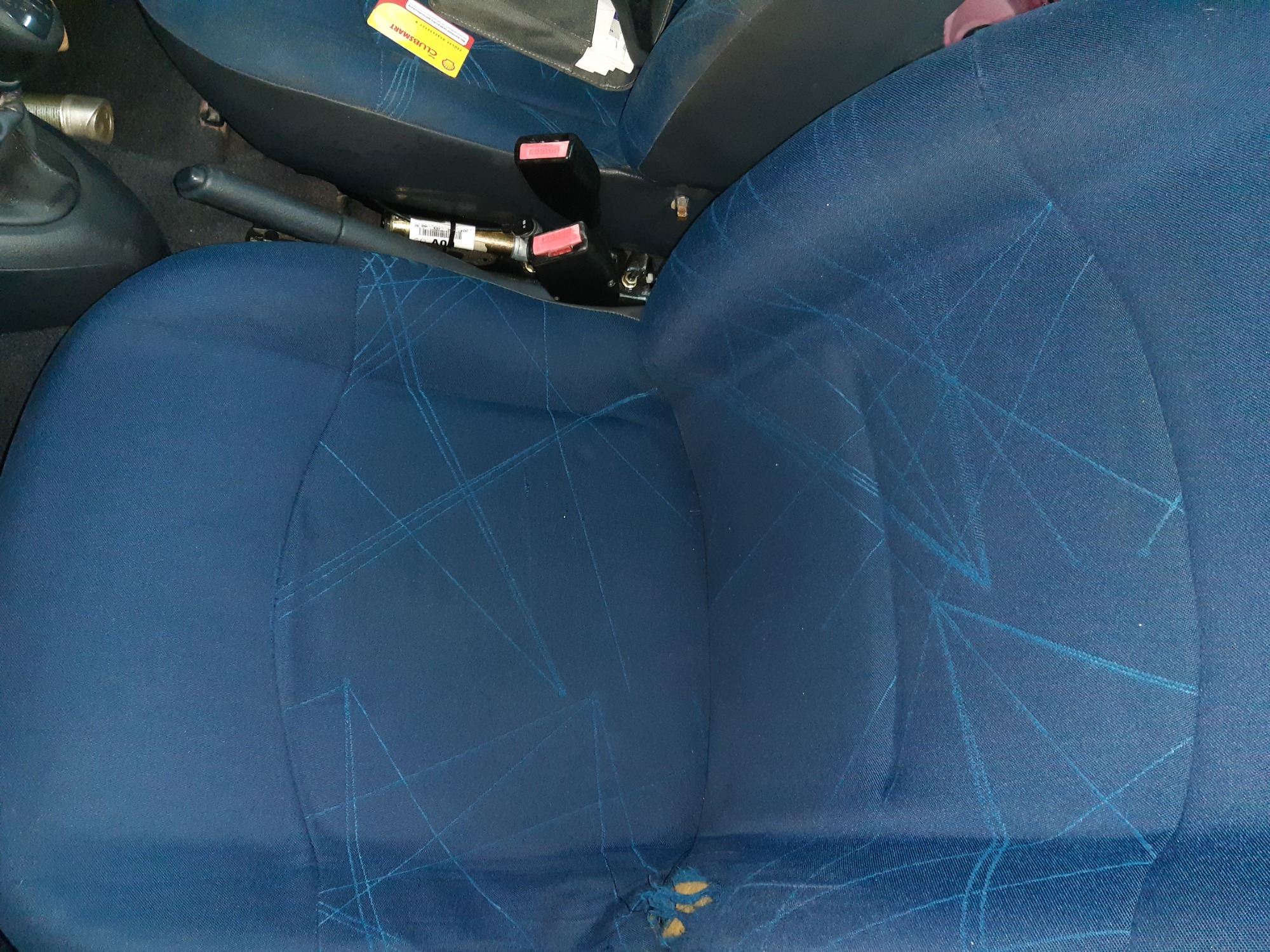 Niebieski fotel samochodowy z dziurą w dolnej części siedziska, widoczny fragment dźwigni hamulca ręcznego i pasy bezpieczeństwa.