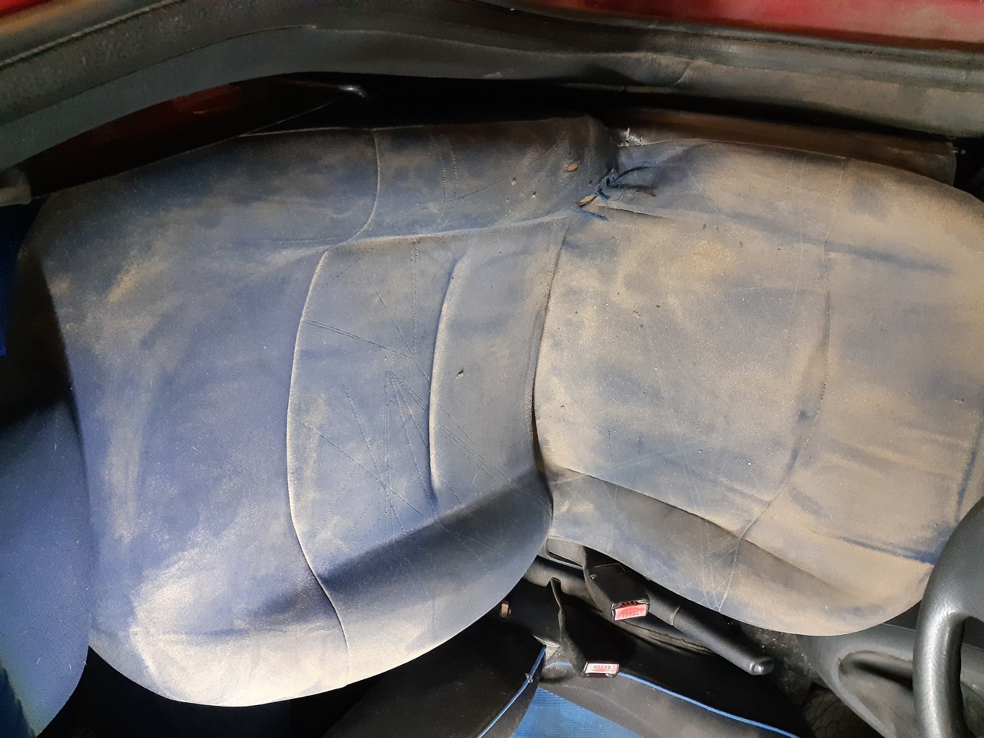 Silnie zabrudzona, błękitna tapicerka tylnej kanapy samochodowej, widoczne zarysowania i plamy, zapięcia pasów bezpieczeństwa.