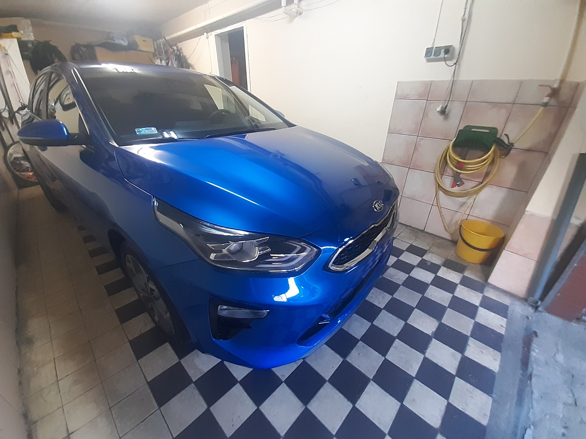 Błękitny samochód Kia zaparkowany w garażu z szachownicową podłogą, widoczny wąż do mycia i żółte wiadro.