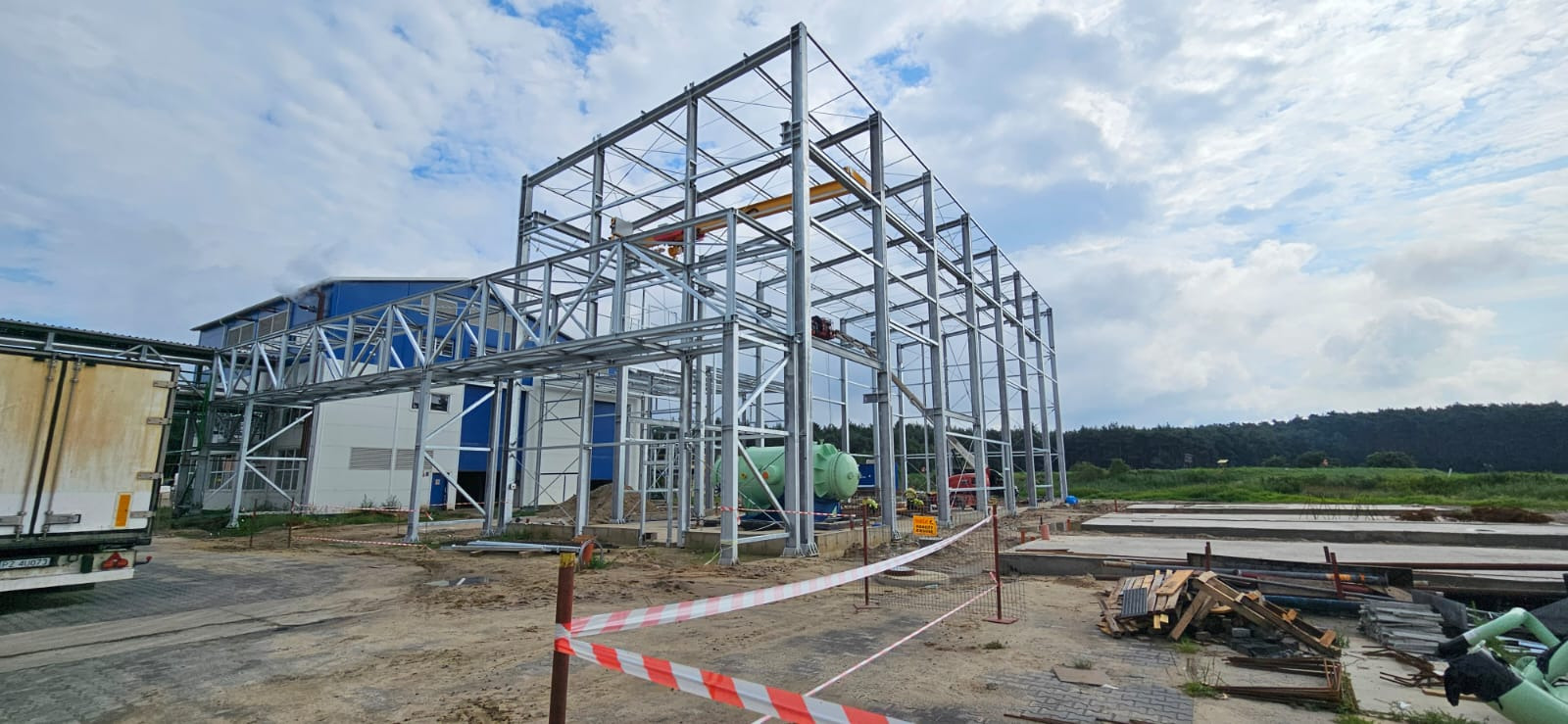 Budowa hali: stalowa konstrukcja szkieletowa, widoczny dźwig, fragment istniejącego budynku, teren budowy z zabezpieczeniami.