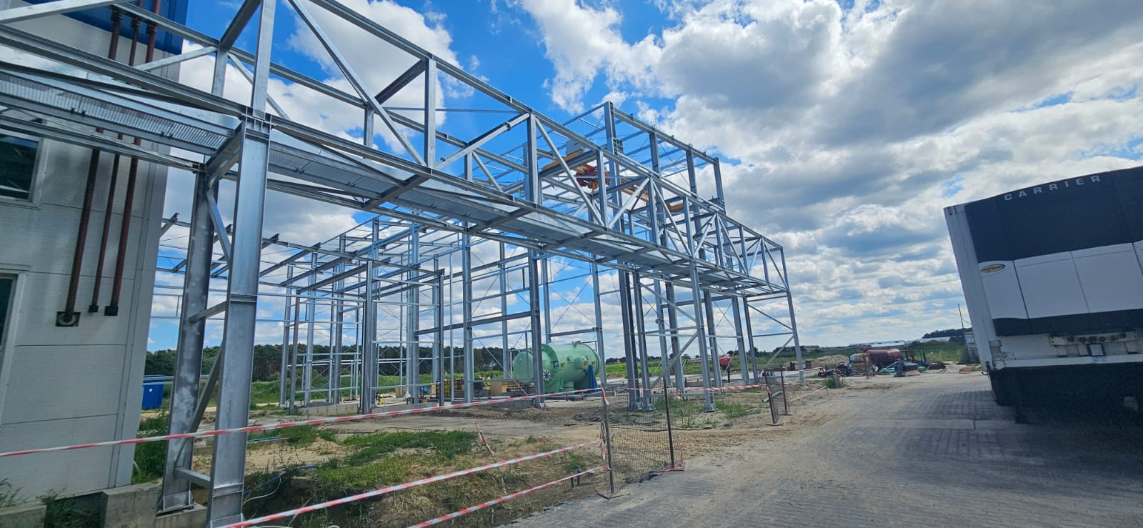 Konstrukcja stalowa hali w trakcie budowy, widok z zewnątrz, błękitne niebo z chmurami w tle. Widoczny fragment budynku z rurami na ścianie i zaparkowana naczepa.