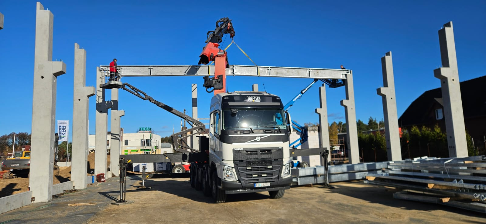 Montaż stalowej konstrukcji hali: żuraw umieszcza belkę na betonowych słupach. Widoczni pracownicy na podnośnikach i na konstrukcji. Transport belek ciężarówką. Słoneczny dzień.