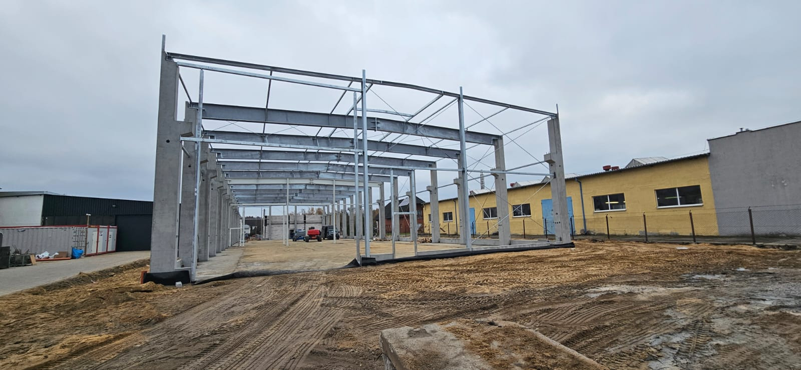 Wznoszenie hali: widok na stalową konstrukcję szkieletową z betonowymi filarami, piaskowe podłoże, sprzęt budowlany w tle, budynek z żółtą elewacją po prawej.