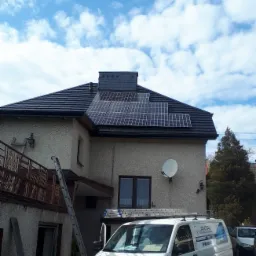 Dom jednorodzinny z panelami słonecznymi na dachu, widoczny samochód dostawczy firmy instalacyjnej z logo i numerem telefonu, drabina oparta o ścianę budynku.