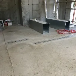 Surowy betonowy fundament w trakcie prac budowlanych, widoczne stalowe pręty zbrojeniowe w podłożu, dwa prostokątne metalowe kanały wentylacyjne, materiały izolacyjne w tle.