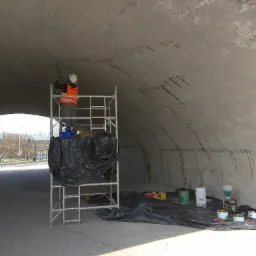 Pracownik w kasku i kamizelce na rusztowaniu, naprawiający betonowy sufit tunelu z widocznymi pęknięciami, używając narzędzia pneumatycznego; na podłodze rozłożona folia ochronna, wiadra...