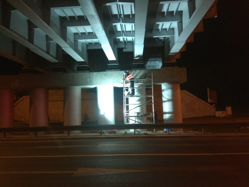 Pracownik w kasku i kamizelce odblaskowej na drabinie, pod betonowym wiaduktem oświetlonym reflektorem, nocą.