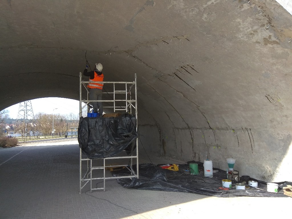 Pracownik w kasku i kamizelce na rusztowaniu, naprawiający betonowy sufit tunelu z widocznymi pęknięciami, używając narzędzia pneumatycznego; na podłodze rozłożona folia ochronna, wiadra...