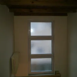 Wnętrze pokoju z oknem z szybami ornamentowymi, drewnianą belką stropową i grzejnikiem żeberkowym przy ścianie.