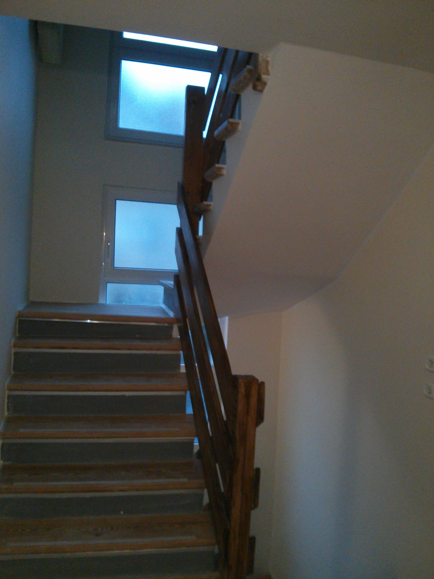 Klatka schodowa w trakcie wykańczania, widoczne schody z drewnianymi elementami, białe ściany i okna z niebieskim światłem w tle.