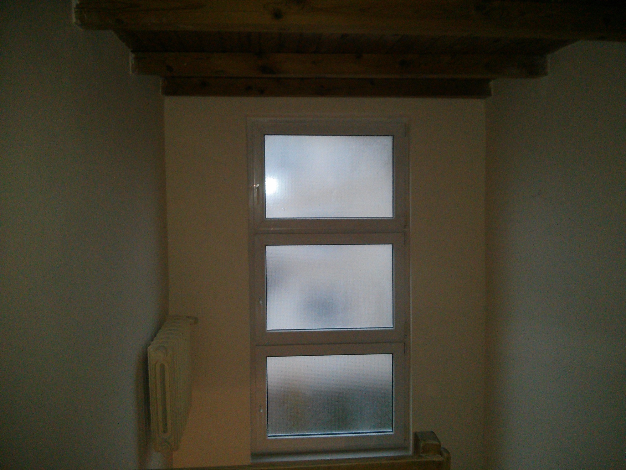 Wnętrze pokoju z oknem z szybami ornamentowymi, drewnianą belką stropową i grzejnikiem żeberkowym przy ścianie.