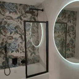 Nowoczesna łazienka z kabiną prysznicową z czarną ramą, deszczownicą i okrągłym lustrem z podświetleniem LED, na ścianie tapeta w kwiaty.