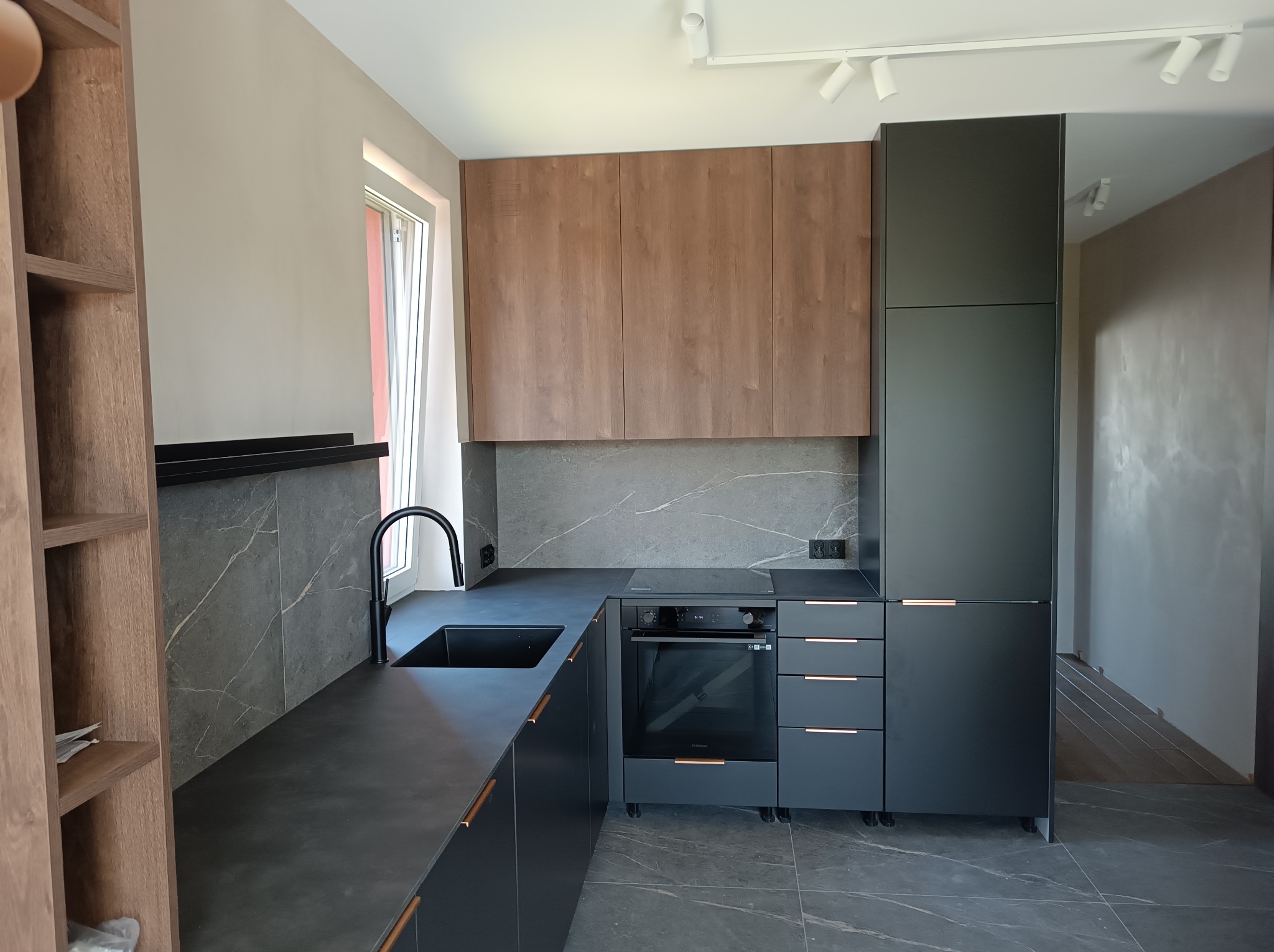 Nowoczesna kuchnia z szarymi marmurkowymi płytkami na ścianie, czarnymi szafkami z miedzianymi uchwytami i drewnianymi szafkami wiszącymi.