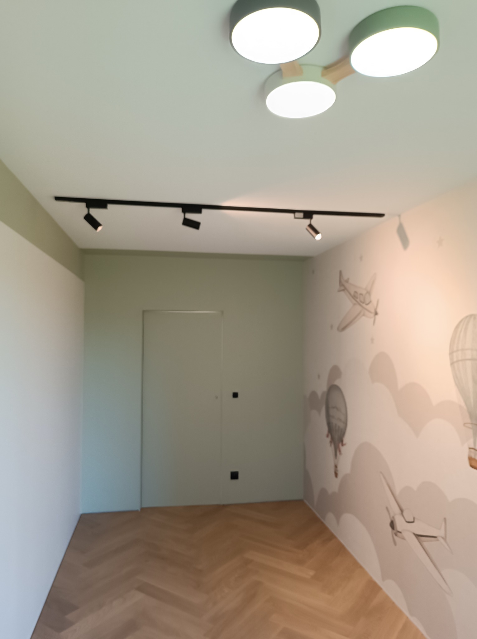 Pokój dziecięcy z jasną podłogą ułożoną w jodełkę, ściany w kolorze złamanej bieli z motywem samolotów i balonów, oświetlenie sufitowe: trzy okrągłe lampy oraz szyna z reflektorami.