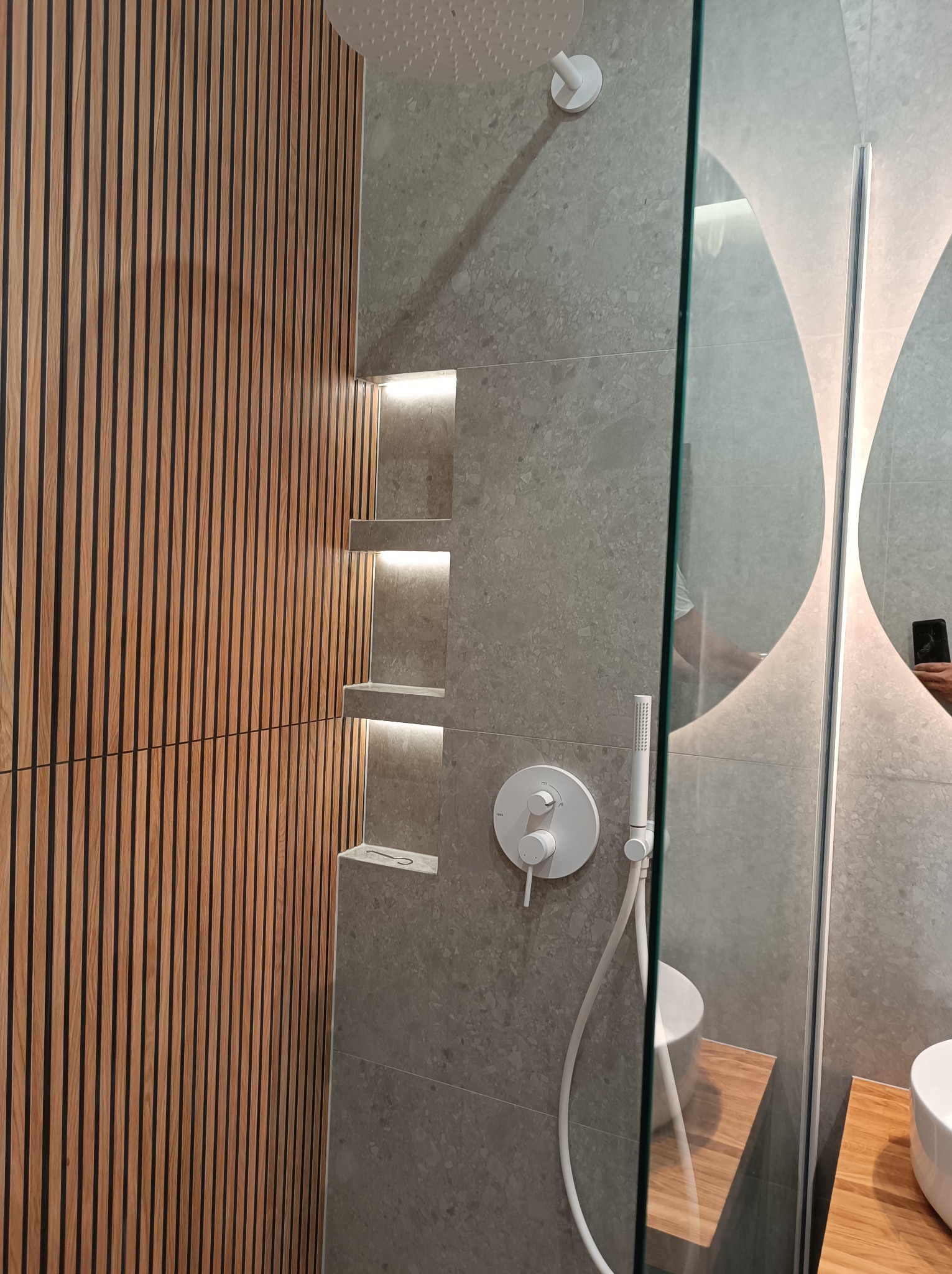 Nowoczesna łazienka z drewnianą ścianą z pionowych listew, szarą płytką imitującą kamień, wnękami z podświetleniem LED, deszczownicą i szklaną kabiną prysznicową.
