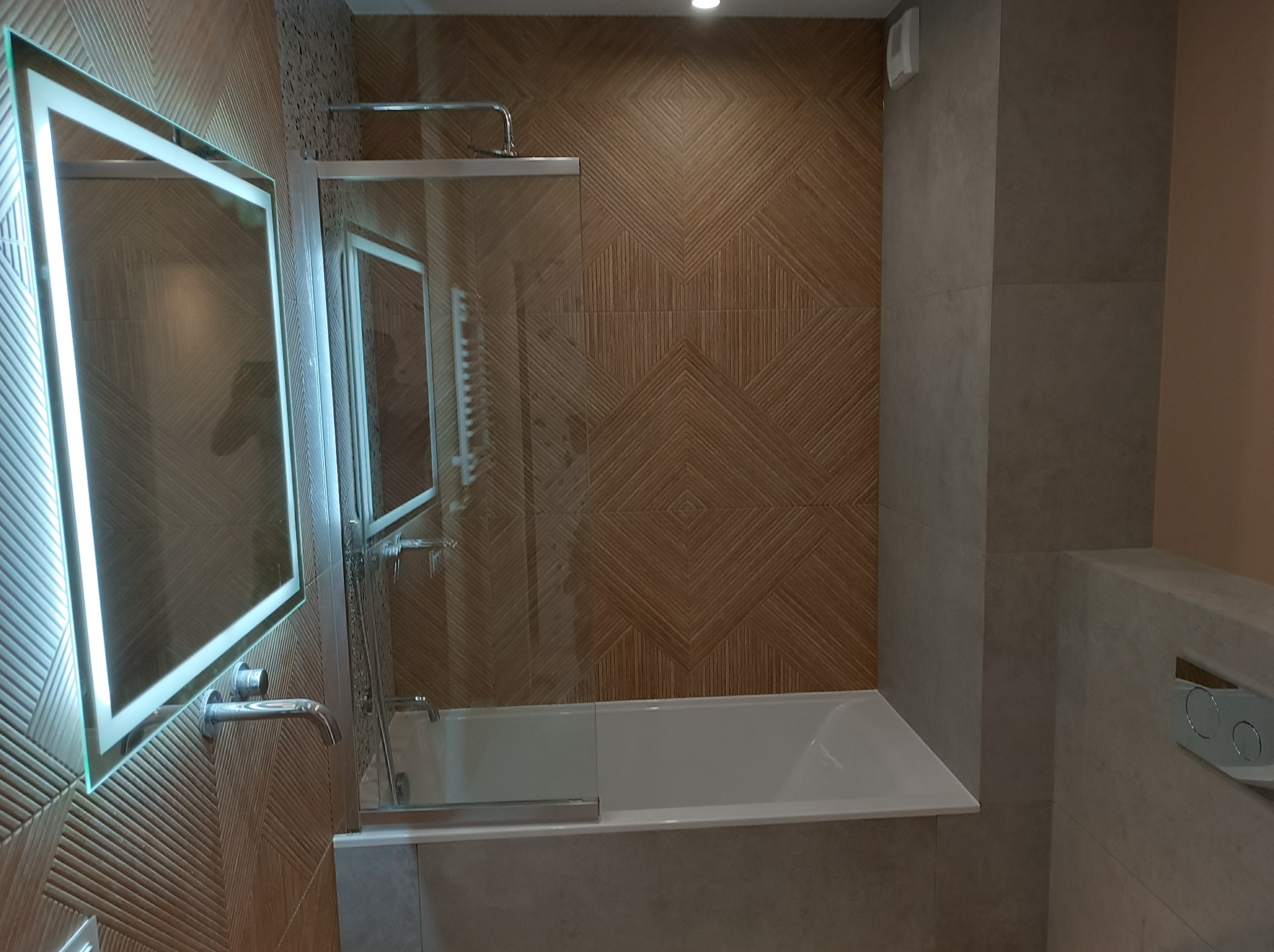 Nowoczesna łazienka z podświetlanym lustrem, kabiną prysznicową ze szklaną ścianką i wanną, wykończona płytkami w odcieniach brązu i szarości z geometrycznym wzorem na ścianie.