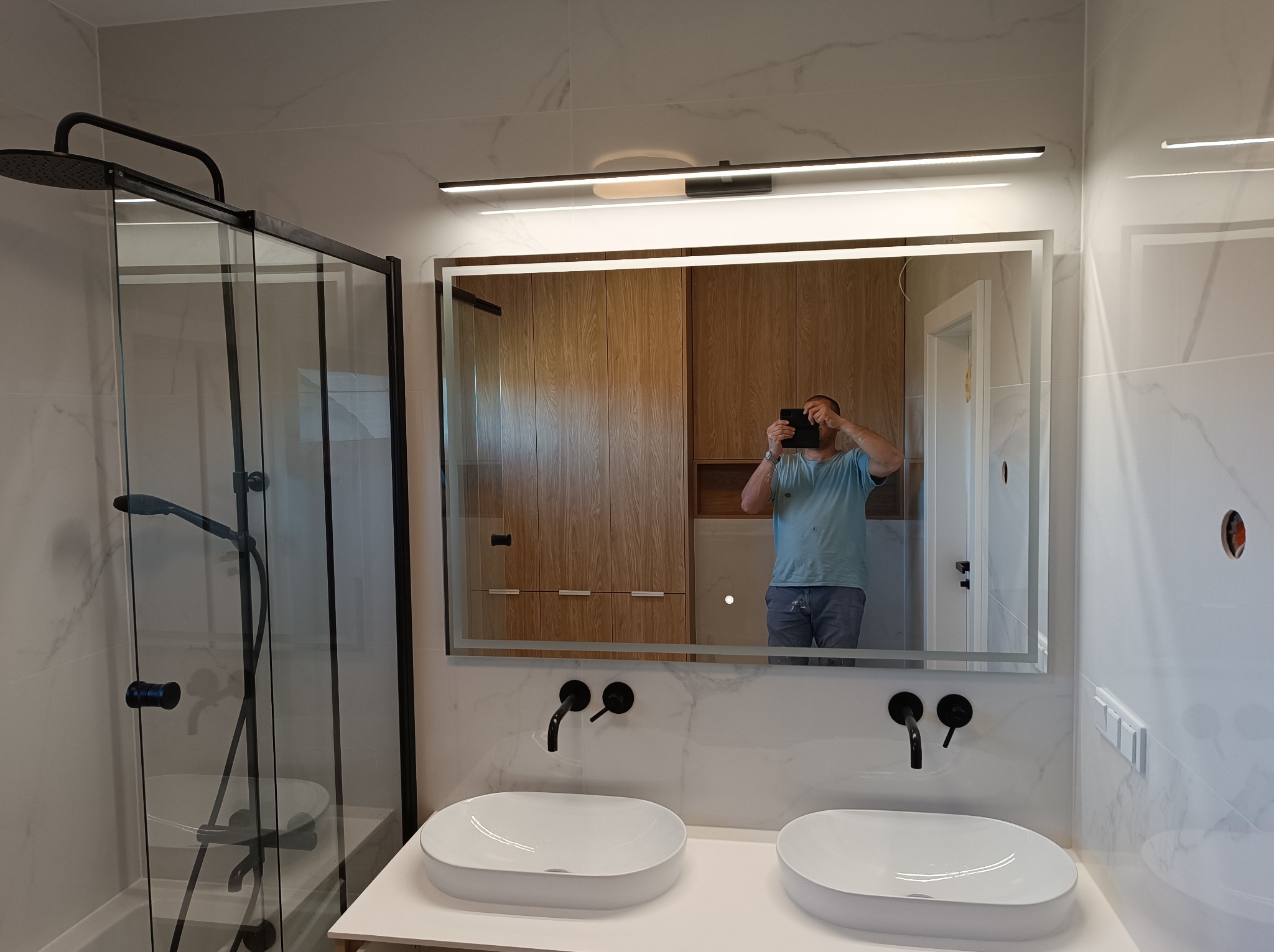 Nowoczesna łazienka z dwoma białymi umywalkami nablatowymi, czarną armaturą i kabiną prysznicową z czarnymi ramami, widok na szafki w odbiciu lustra.