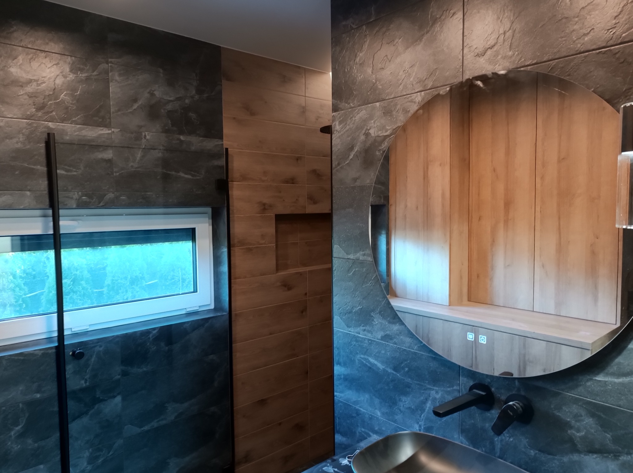 Nowoczesna łazienka z płytkami imitującymi marmur i drewno, okrągłe lustro z podświetleniem LED, czarna armatura podtynkowa i okno w kabinie prysznicowej.