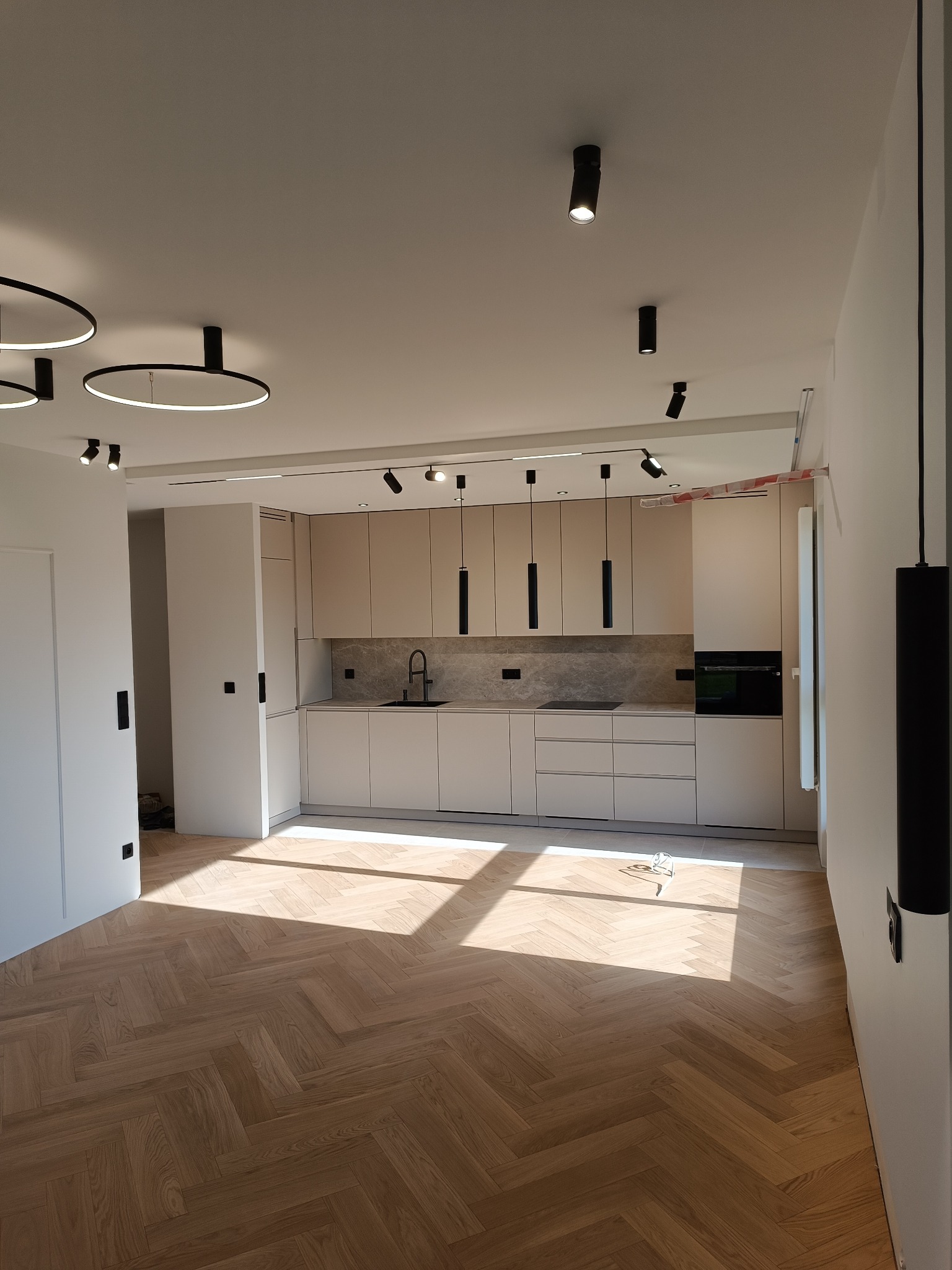 Nowoczesna kuchnia z jasnymi szafkami, szarym blatem i parkietem w jodełkę, oświetlona promieniami słońca wpadającymi przez okno.