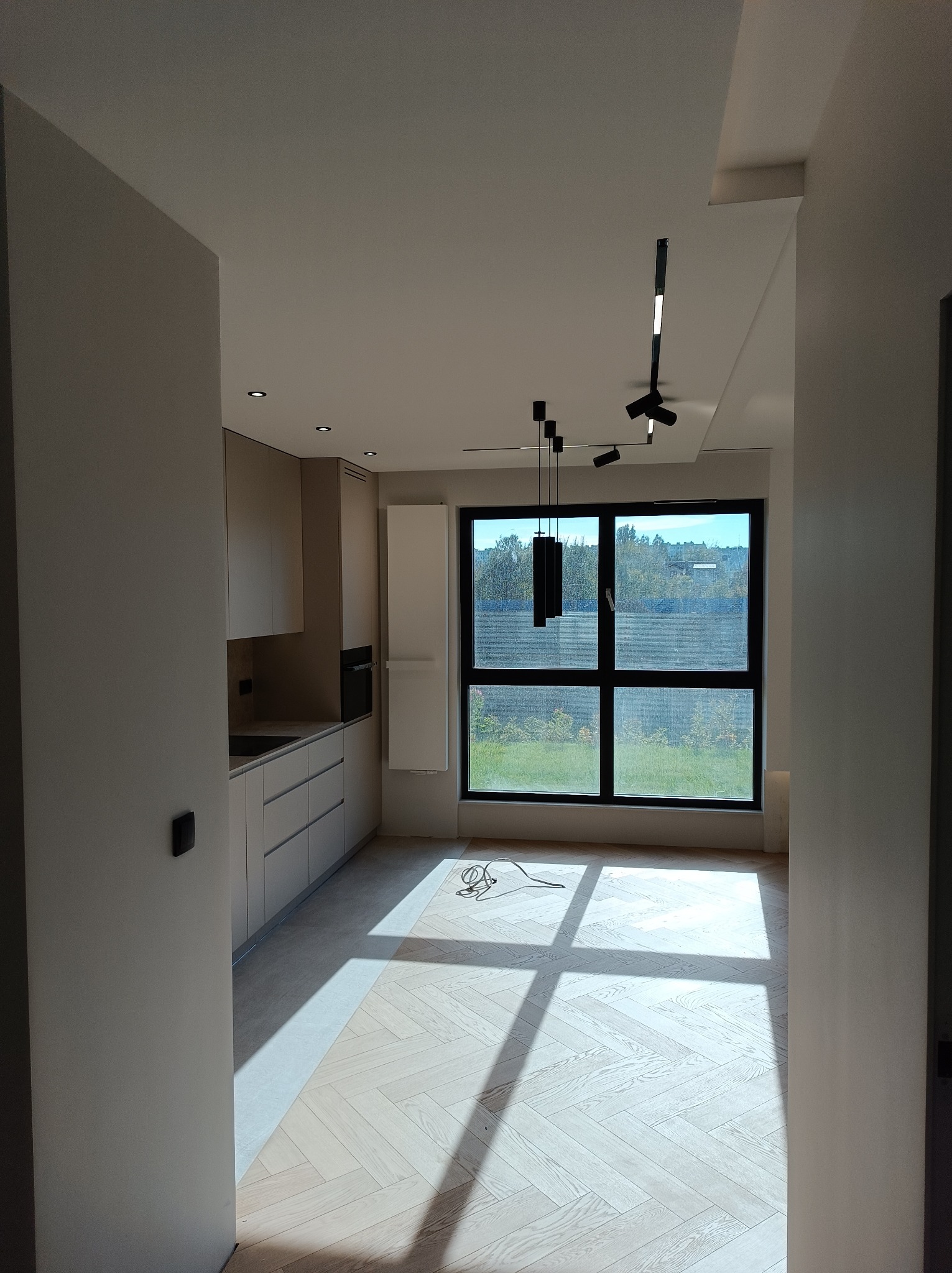 Jasne, przestronne wnętrze w trakcie wykańczania: widok na kuchnię z beżowymi szafkami, parkiet ułożony w jodełkę, duże okno z widokiem na zieleń i nowoczesne czarne lampy sufitowe.