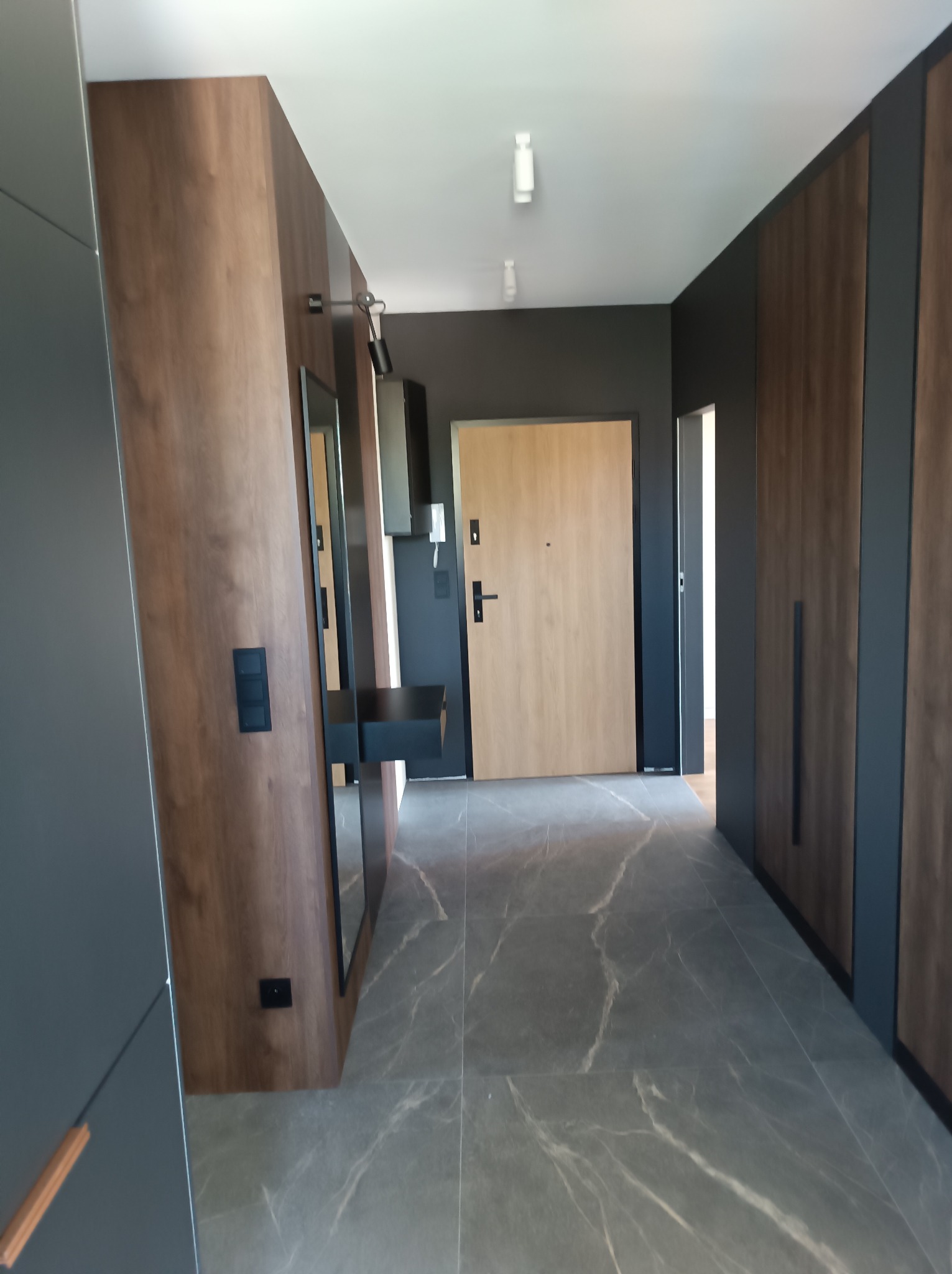 Nowoczesny przedpokój z szarą marmurową podłogą, drewnianymi panelami ściennymi i szafą w zabudowie z ciemnymi elementami wykończeniowymi, lustro z czarną konsolą, drzwi wejściowe w kolorze jasnego...