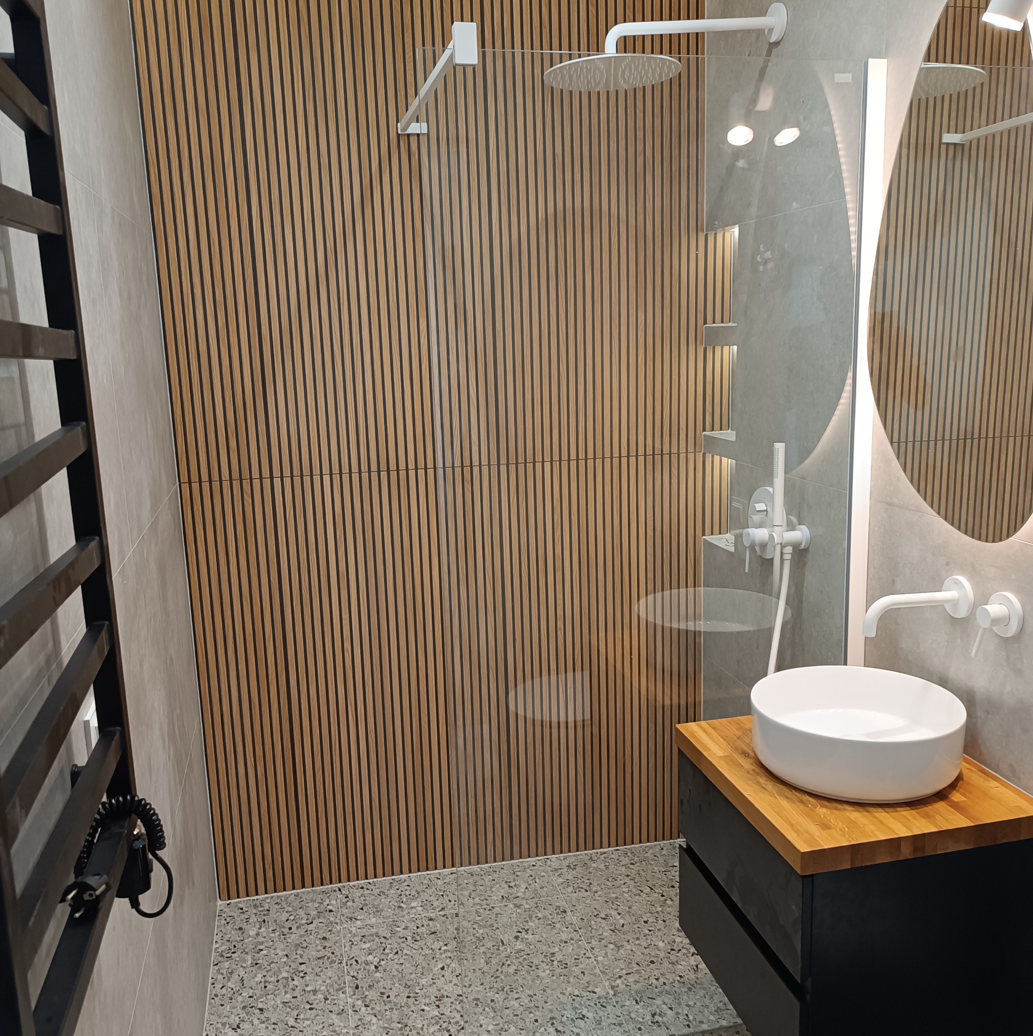 Nowoczesna łazienka z drewnianymi lamelami na ścianie prysznica, okrągłym lustrem z podświetleniem LED, białą umywalką nablatową na drewnianym blacie i grzejnikiem drabinkowym.