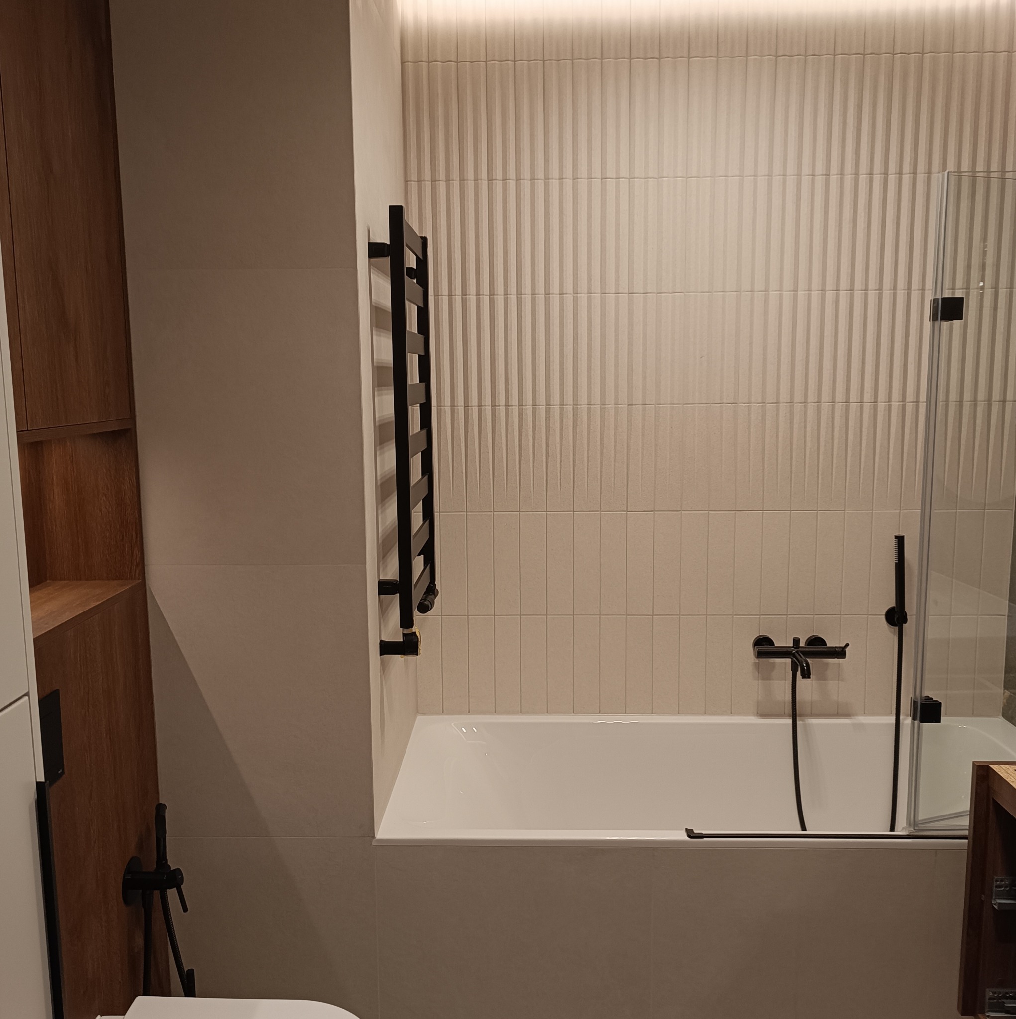 Nowoczesna łazienka z wanną obudowaną płytkami, szklanym parawanem nawannowym, grzejnikiem drabinkowym w kolorze czarnym i armaturą w tym samym kolorze.