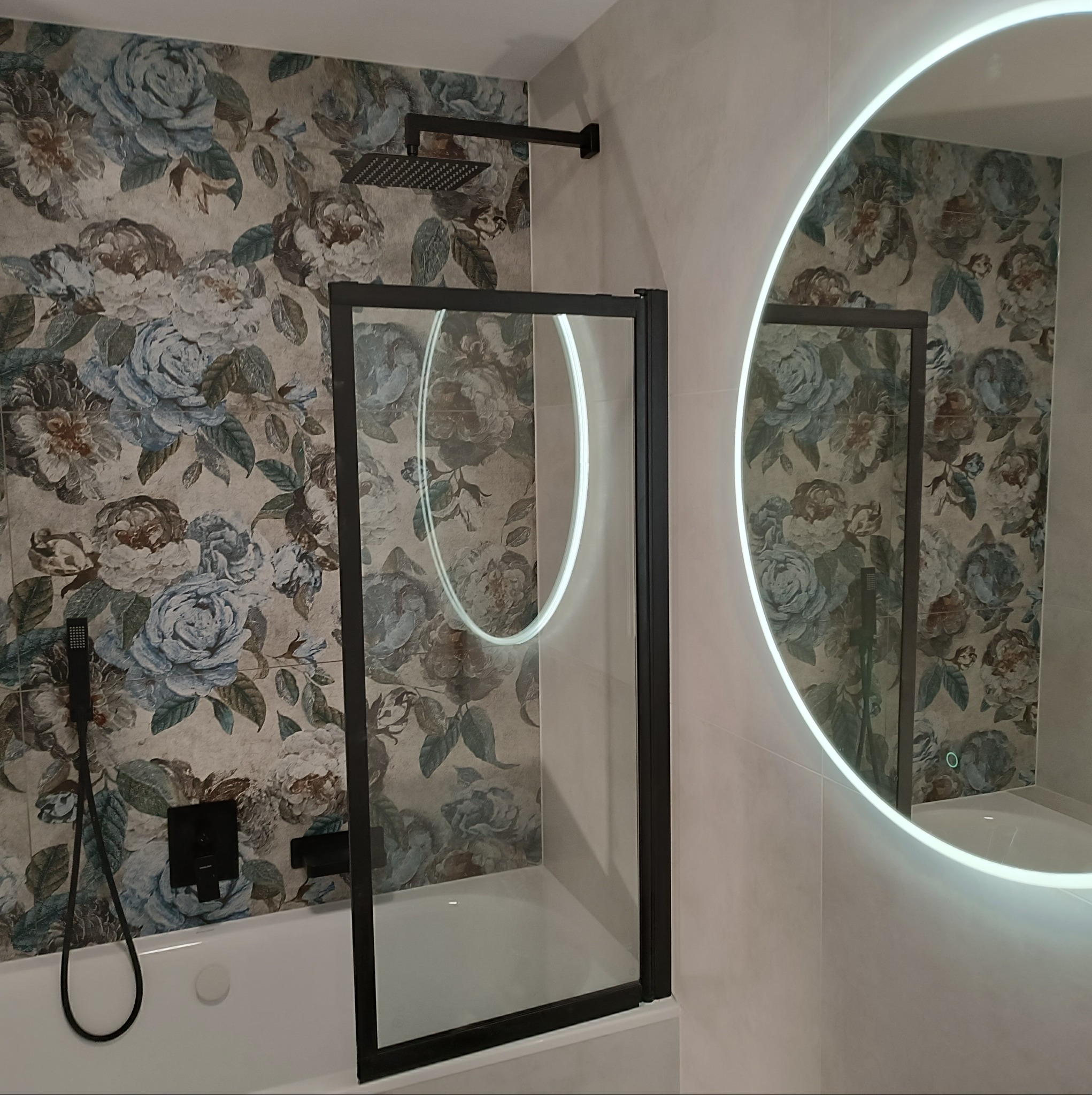 Nowoczesna łazienka z kabiną prysznicową z czarną ramą, deszczownicą i okrągłym lustrem z podświetleniem LED, na ścianie tapeta w kwiaty.