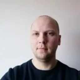 Portret mężczyzny o jasnej karnacji, z krótkimi włosami, w ciemnej bluzie na tle jasnej ściany. Ujęcie od przodu, skupione na twarzy i górnej części tułowia.