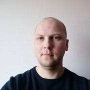 Portret mężczyzny o jasnej karnacji, z krótkimi włosami, w ciemnej bluzie na tle jasnej ściany. Ujęcie od przodu, skupione na twarzy i górnej części tułowia.