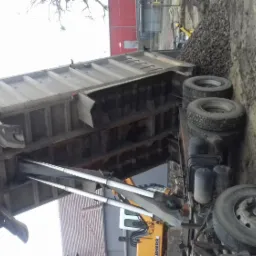 Ciężarówka wywrotka z podniesioną skrzynią ładunkową, wysypująca ciemny żwir na ziemię. W tle widoczna koparka i budynek.