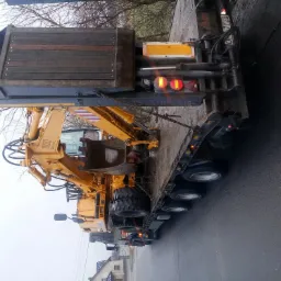 Żółta koparka Liebherr 900 Litronic przewożona na niskopodwoziowej naczepie ciężarówki z włączonymi światłami pozycyjnymi, widok z tyłu na tle szarej drogi i drzew bez liści.