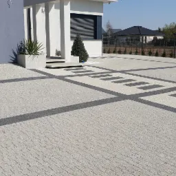 Nowoczesny podjazd z jasnoszarej kostki brukowej z ciemnoszarymi wstawkami, prowadzący do domu z szarą bramą garażową i białymi kolumnami przy wejściu, z widocznym fragmentem białego samochodu.