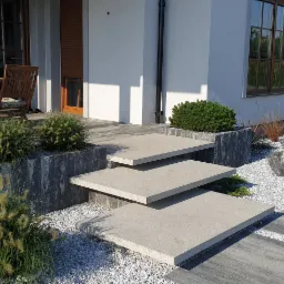 Nowoczesne schody wejściowe z jasnego betonu prowadzące do domu, otoczone ozdobnymi kamieniami, trawami i niskimi krzewami, z widocznymi fragmentami tarasu i mebli ogrodowych w tle.