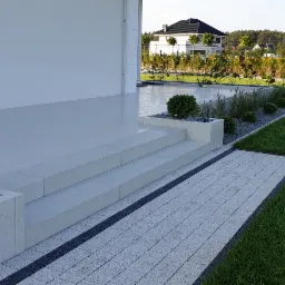 Nowoczesny taras z jasnych płyt, dwustopniowe schody z donicami z kamieniami i kulistymi tujami, obok trawnik i rabaty z ozdobnymi trawami, w tle dom jednorodzinny.